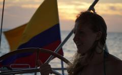 A woman smiles, steering a boat with a Colombian flag waving in the background at sunset.