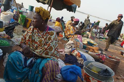 A woman in colorful attire sits at a market stall, surrounded by goods, while other vendors and customers mill about in the background.