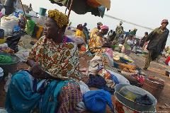 A woman in colorful attire sits at a market stall, surrounded by goods, while other vendors and customers mill about in the background.