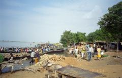 A bustling market scene unfolds by a river with boats, people, and a donkey loaded with goods under a cloudy sky.