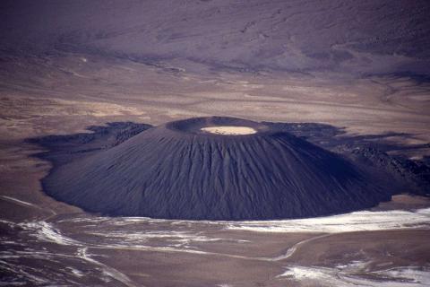 The Quest of the Tibesti Mountains An aerial view of a volcanic cone in the Tibesti Mountains, with a crater at the summit and surrounding barren, dusty landscape.