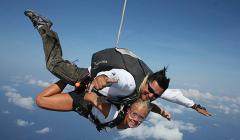 Um homem e uma mulher estão praticando paraquedismo, presos um ao outro, com a mulher sorrindo e fazendo sinal de positivo, tendo como pano de fundo o céu e as nuvens.