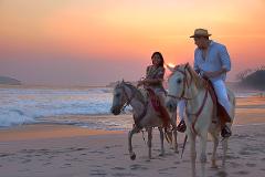A couple rides horses on a beach at sunset in Nicaragua, with the golden sky reflecting on the sand and waves.