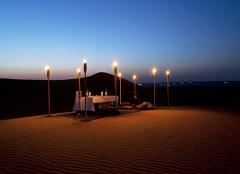 A romantic dinner table set up in the desert at dusk, illuminated by tiki torches, offers a magical experience under the twilight sky.