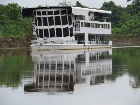 A white houseboat named "Sepik Spirit" is floating on a calm river, reflecting its image on the water, with lush green vegetation on the riverbanks.