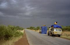 Two off-road vehicles are parked on a long, straight road, and two men secure a blue tarp over cargo on the roof, under a dark, stormy sky.