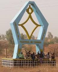 A blue monument with the words "Festival de Tabiaskia" inscribed on it, with a group of people sitting at its base, against a backdrop of a rural landscape.