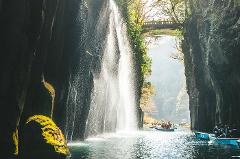 日本の高千穂峡の美しい景色。川に流れ落ちる滝と、ボートを楽しむ観光客が描かれており、背景には橋が見えます。