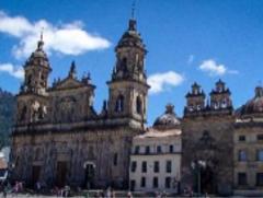 A view of the Metropolitan Cathedral Basilica of Bogotá, a large stone cathedral with towers, domes, and intricate carvings, set against a blue sky.