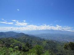A scenic view of green mountains under a partly cloudy blue sky, with a city visible in the distance.