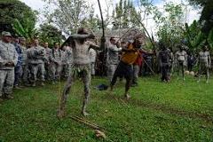 Certainly! Here's a description of the image within the character limit:A group of US military personnel observes a cultural demonstration, likely in Papua New Guinea, featuring men in traditional Asaro Mudmen attire engaging in a ritual performance.