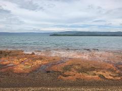 A serene landscape of a beach with rusty-colored rocks leading to a calm sea under a cloudy sky.