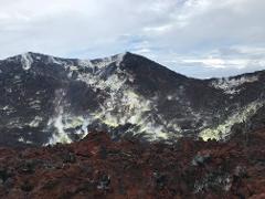 A mountainous volcanic landscape features dark rock formations with yellow deposits and steam vents under a cloudy sky.