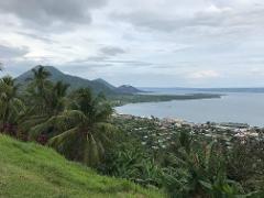 A scenic view of Rabaul, Papua New Guinea, featuring lush green hills, palm trees, a coastline with a bay and mountains in the background under a cloudy sky.