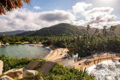 Tayrona National Park Private Luxury Tour (3 Days / 2 Nights) A secluded tropical beach with lush green hills and palm trees, a small river flowing into the ocean, and a few people enjoying the sun and water.