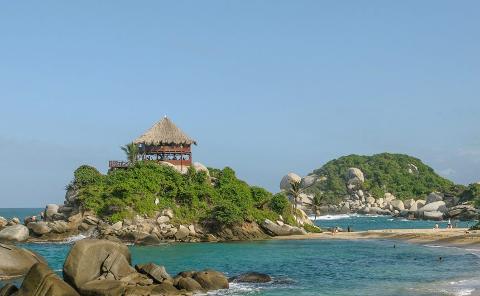 Tayrona National Park Private Luxury Tour (3 Days / 2 Nights) A thatched-roof hut sits atop a rocky, verdant island in Tayrona National Park, with turquoise waters and a sandy beach dotted with people.