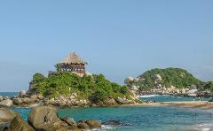 Tayrona National Park Private Luxury Tour (3 Days / 2 Nights) A thatched-roof hut sits atop a rocky, verdant island in Tayrona National Park, with turquoise waters and a sandy beach dotted with people.
