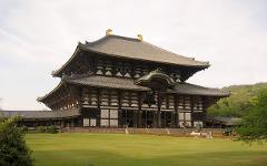 A large, multi-tiered wooden temple with a dark tiled roof and golden ornaments stands prominently in a green grassy area, with people visible near the entrance.