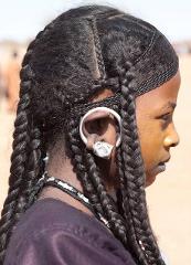 A close-up side profile of a young girl with braided hair, a large silver earring, and a beaded necklace, set against a desert background.