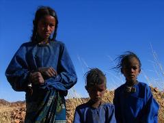 Three children, possibly from a nomadic tribe, stand against a clear blue sky in a desert environment, dressed in traditional blue clothing and necklaces.