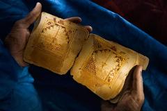 A person's hands hold open an old, worn book with decorative borders and intricate designs, set against a textured blue cloth background.