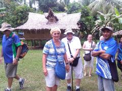 A group of tourists, including a man with crossed arms in the foreground, pose in front of a traditional thatched-roof hut in Alotau, Milne Bay, surrounded by lush tropical greenery.