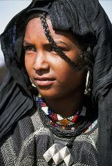 A young woman with dark skin and a black head covering, adorned with a single braid, wears colorful beaded necklaces and a silver pendant against a textured dark fabric.