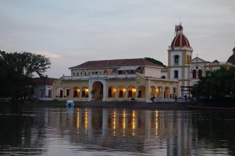 Mompox & Aracataca Cultural Journey (3 Days / 2 Nights) A yellow colonial-style building with a red-domed church sits on the bank of a reflective river at dusk.