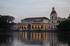 Mompox & Aracataca Cultural Journey (3 Days / 2 Nights) A yellow colonial-style building with a red-domed church sits on the bank of a reflective river at dusk.