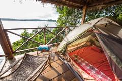 A tent is set up on a wooden deck overlooking a serene lake, with comfortable seating and lush greenery.