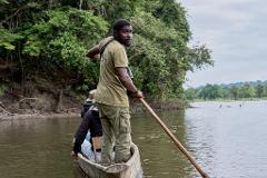 A man in a canoe navigates a river bordered by dense forest, looking back at the camera with a serious expression, while others sit behind him in the boat.