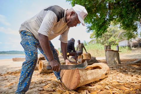 An African man carves a log with a machete, with two other men and a river in the background.