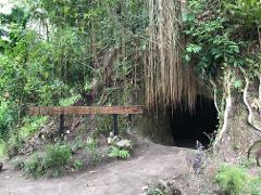 A cave entrance framed by lush vegetation and a wooden sign reading "Project Funded by National Gaming Control Board" in Papua New Guinea.