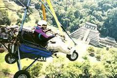 Here's a description of the image:An ultralight aircraft with a pilot is flying in front of an ancient pyramid and lush green scenery.