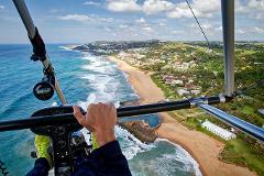 This aerial shot captures a scenic coastline from an ultralight aircraft, with a person holding the controls and vibrant ocean waves crashing onto the sandy beach.