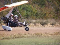 An ultralight aircraft with two pilots wearing helmets is on a dirt road with trees in the background.