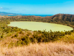 A scenic view of a green-colored lake surrounded by lush greenery, with a blue sky and distant land in the background, captured from a grassy hilltop.