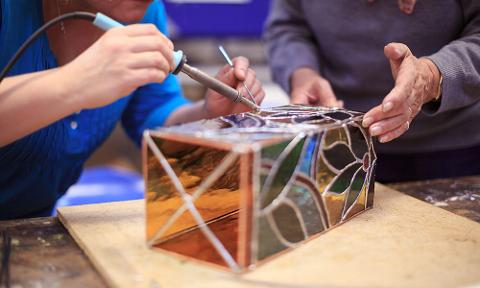 Two people are crafting a stained glass box, one soldering and the other holding it in place.