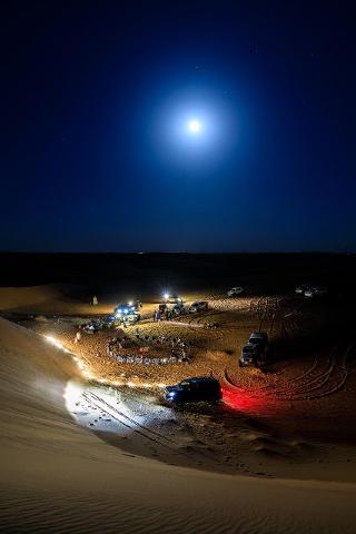 A group of people gather in the desert under a bright moon, surrounded by cars and a circle of chairs, with tire tracks in the sand.