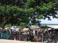 Et travlt udendørsmarked i Wewak, Papua Ny Guinea, med sælgere og kunder under parasoller og et stort træ, med udsigt over havet i baggrunden.