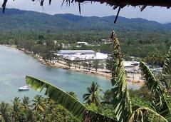 Wewak Day Tour Here's a description of the image:A scenic view of Wewak coastline features a bay, a white building complex along the shore, lush green mountains in the background, and tropical foliage in the foreground, creating a vibrant tropical landscape.