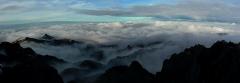 Panoramatický pohľad z vrcholu hory Mount Wilhelm ukazuje rozoklané štíty prenikajúce cez more oblakov pod modrou a zamračenou oblohou.
