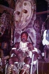 Certainly! Here is a description of the image within the character limit:A man dressed in ceremonial attire sits in front of a large, elaborate wooden mask, likely a part of the Sepik River Crocodile Festival.