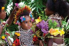 A group of elaborately decorated children are shown in traditional attire, including colorful feathers, shell necklaces, and painted faces, likely participating in a cultural event in Papua New Guinea.
