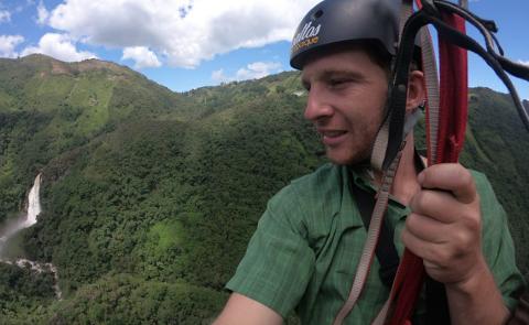 A man is paragliding over a lush green landscape with a waterfall visible in the distance.