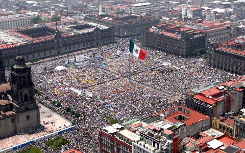 Aerial view of a densely packed crowd filling a vast plaza in Mexico City, surrounding a large Mexican flag, with historic buildings in the background.