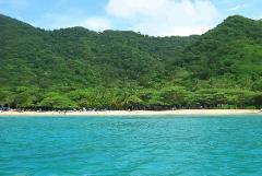Tayrona Park & Playa Cristal Full Day Tour A scenic beach with turquoise water meets a sandy shore lined with trees, backed by lush green hills.