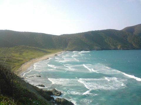 Tayrona Park & Playa Cristal Full Day Tour A scenic view of a beach with crashing waves, a sandy shoreline, and lush green hills in the background under a bright sky.