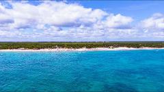 Tayrona Park & Playa Cristal Full Day Tour A scenic aerial view shows a turquoise ocean meeting a white sandy beach lined with lush green trees under a bright blue sky filled with fluffy white clouds.
