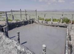 Private Cartagena Mud Volcano + Mangrove Tour including lunch A gray pool of mud is surrounded by a makeshift wooden and concrete barrier, with a murky green lake and distant mountains in the background under a partly cloudy sky.